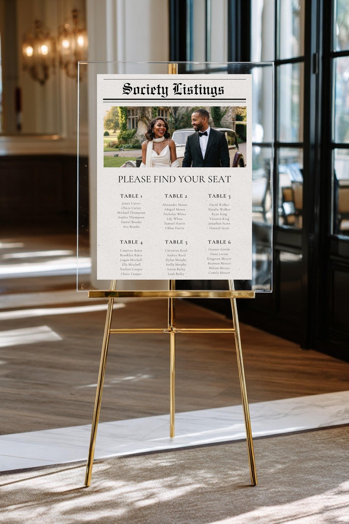 Newspaper-style wedding seating chart poster on gold easel, with photo header and table list